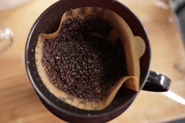 Detaylı bir yukarıdan çekim, açık kahverengi bir kağıt filtreye yerleştirilmiş ıslak, kavrulmuş kahve tellerini yakalar ve sıcak ahşap bir yüzeyde, siyah bir kahve damlatıcısının içine yerleştirir. Bu resim yeni bir bardak dri 'nin temel bileşenlerini gösteriyor