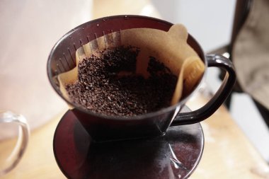 Yakından bakıldığında koyu, kaba kahveyi, açık kahverengi bir kağıt filtreye dikkatlice yerleştirilmiş, siyah bir kahve damlatıcısının içine yerleştirilmiştir. Bu kurulum taze, aromatik kahve demlemek için hazırlandı, basit ama gerekli araçları vurguluyor.