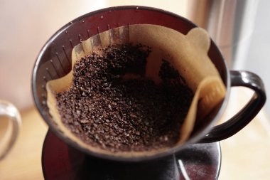 Detaylı bir çekim koyu kavrulmuş, kahverengi bir kağıt filtrede koyu kahve damlatıcısının içinde kabaca öğütülmüş kahve çekirdeklerini gösteriyor. Kurulum elle demlenmeye hazır, zeminin dokusunu vurguluyor ve temel malzemeler hazırlanıyor..