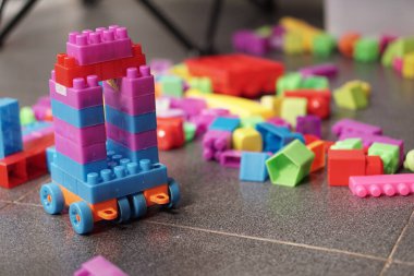 Pembe, kırmızı ve mavi birbirine kenetlenmiş plastik yapı taşlarından yapılmış canlı bir oyuncak araç koyu kiremitli bir zeminde belirgin şekilde duruyor. Diğer renkli plastik tuğlalar yumuşak bulanık arkaplana saçılmış.