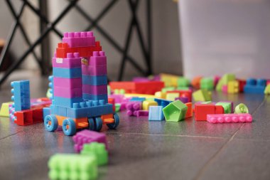 Yakın plan bir görüntü, fayanslarla döşenmiş bir zemine yayılmış canlı bir dizi plastik yapı taşını yakalar. Farklı renklerde birbirine kenetlenmiş bloklardan yapılmış ve tekerleklerle donatılmış yaratıcı bir oyuncak araç, oyuncu benliğimin ortasında duruyor.