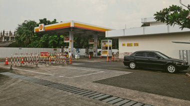 Jakarta - Endonezya, 02 / 03 / 2026: Bulutlu bir gökyüzü altında çoklu yakıt pompaları, yakınlarda park edilmiş siyah bir sedan ve ön planda kırmızı ve beyaz güvenlik bariyerleri bulunan bir Shell V-Power benzin istasyonu gösteriliyor.