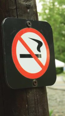 Ağaç gövdesine yapıştırılmış kırmızı çizgi ve daire şeklinde sigara içilmez işareti olan isim levhası. Arka plandaki eğlence parkı aile dostu açık hava mekanının dikey çekim yaptığını gösteriyor.