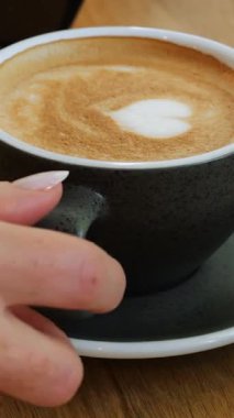 Zarif kadın eli, siyah seramik kahve fincanını ahşap masadan alır. Bayan misafir, kahve dükkanında dikey videoda kalp dekoru olan kupadan lezzetli bir kapuçino içiyor.