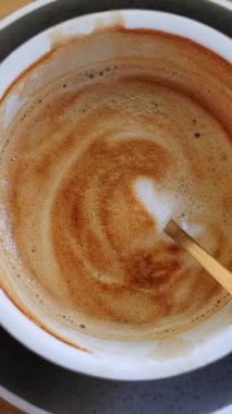 Köpüklü kremalı latte altın çay kaşığı ve ahşap tezgahın üzerinde porselen fincan. Cafe dikey videoda seramik bardaktan köpükle taze kahve içmek.