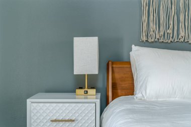 Modern yatak odası içinde beyaz pirinç lamba, komodin, yatak, yastık ve makrome duvar sanatı var.