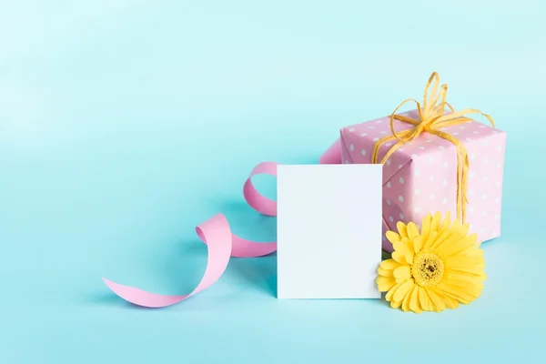 Caja de regalo punteada rosa, flor de gerberas amarillas y tarjeta vacía sobre un fondo azul . — Foto de Stock