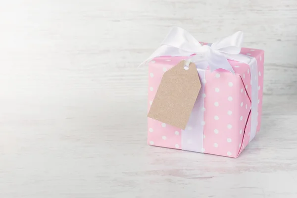 Ajándék doboz papírba csomagolva rózsaszín pontozott, kötött szatén íj és egy üres kraft kártya, mint egy fehér fa háttér. — Stock Fotó