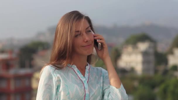 Красивая молодая профессиональная женщина имеет телефонный разговор на крыше офиса балкон в городе в течение дня. Она стоит по пояс, улыбается и волосы развиваются на ветру. Катманду, Непал — стоковое видео