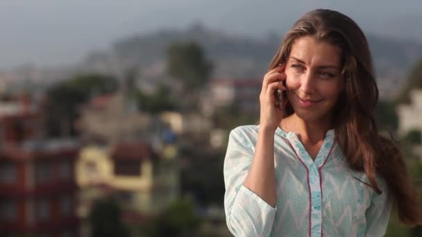 Hermosa joven profesional tiene una conversación telefónica en el tejado de la oficina balcón en la ciudad durante el día. Ella se pone de pie hasta la cintura, sonriendo y el pelo se desarrolla en un viento. Katmandú, Nepal — Vídeo de stock