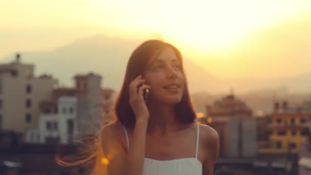 Стільниковий телефон молода жінка говорити на смартфон Sun City Sunset. Молодий спеціаліст, посміхаючись, сміючись, маючи розмову на мобільний телефон волосся розвіваються вітер. Літо, Катманду, Непал. — стокове відео