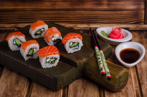 Rollos de Sushi con salmón y pepino servidos sobre tabla de madera con sause de soja y forma de jengibre rosa sobre fondo de madera. Enfoque selectivo —  Fotos de Stock