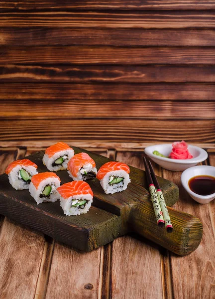 Rollos de Sushi con salmón y pepino servidos sobre tabla de madera con sause de soja y forma de jengibre rosa sobre fondo de madera. Enfoque selectivo —  Fotos de Stock