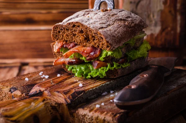 Burger sandwich with lettuce, roasted bacon on dark wood cutting board. Selective focus — Stock Photo, Image