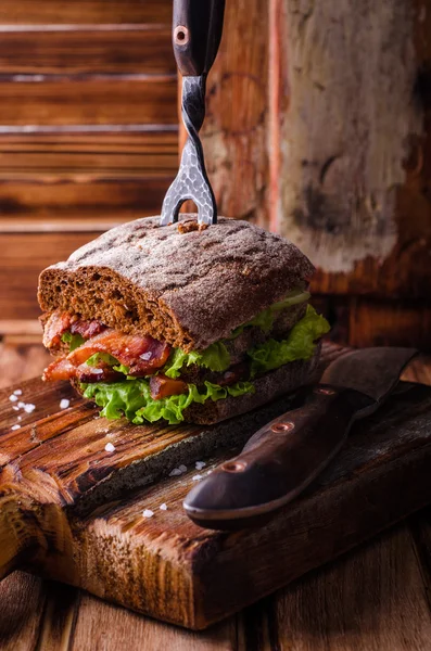 Burger sandwich with lettuce, roasted bacon on dark wood cutting board. Selective focus — Stock Photo, Image