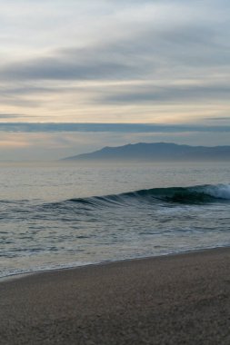 Kumlu bir sahilde dalgalar kırılıyor. Sprey, beyaz su ve çimenler ön planda.
