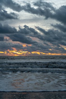 Kumlu sahil şeridi, ağır bulutlar ve gün batımında gelen dalgalar.