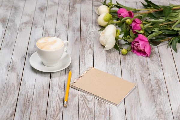 Notebook z ołówka obok kawy i piwonie kwiaty na podłoże drewniane. — Zdjęcie stockowe