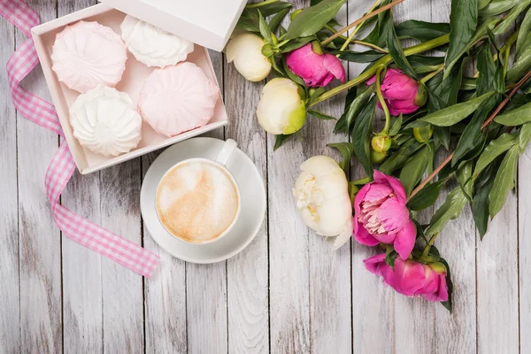 A cup of coffee next to bouquet of peonies, a box with marshmallows and pink ribbon on wooden background. Top view.