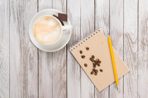 Ноутбук с карандашом рядом с кофе и кофейными зёрнами, кусочек шоколада на белом деревянном фоне. Вид сверху . — стоковое фото