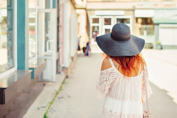 Vista trasera de una chica con el pelo rojo en un sombrero negro caminando por la ciudad. Día soleado de verano. Ciudad style.Girl usando vestido de verano elegante de encaje . — Foto de Stock