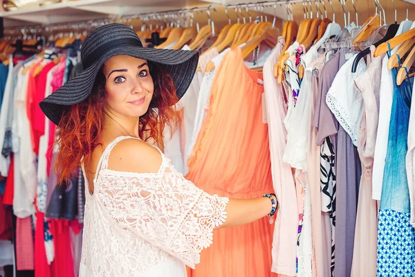 Mladá krásná žena v černém klobouku se snaží najít nové oblečení v obchodě. Při pohledu na fotoaparát. Čas na nákupy. Letní výprodej. — Stock fotografie