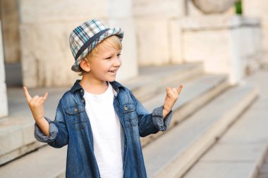 Heavy metal tabelasını tasvir eden bir çocuk. Çocukluk yaşam tarzı konsepti. Kot ceketli ve şapkalı çocuk. Şık küçük çocuk şehirde yürüyüşe çıkmış..