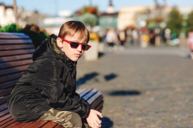 Şehirdeki bankta oturan tatlı çocuk. Siyah ceketli ve son moda güneş gözlüklü yakışıklı çocuk. Şık kış çocukları kıyafeti. Moda, yaşam tarzı ve çocukluk konsepti.