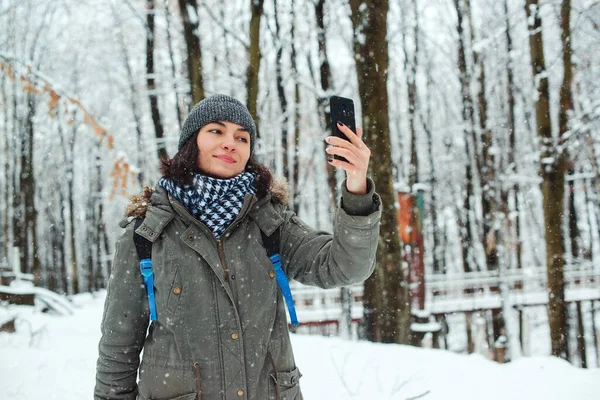 Woman enjoying first snow. Girl walking in winter forest.Woman taking selfie by smartphone in winter. People, technology and season concept.Happy girl enjoying life during snowfall outdoors.