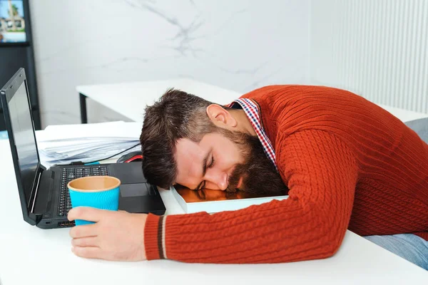 Tired businessman sleeping in office. Bearded man sleeping at his working place. Young business man sleep on the office desk with coffee in hand. Bearded man overworked at office.