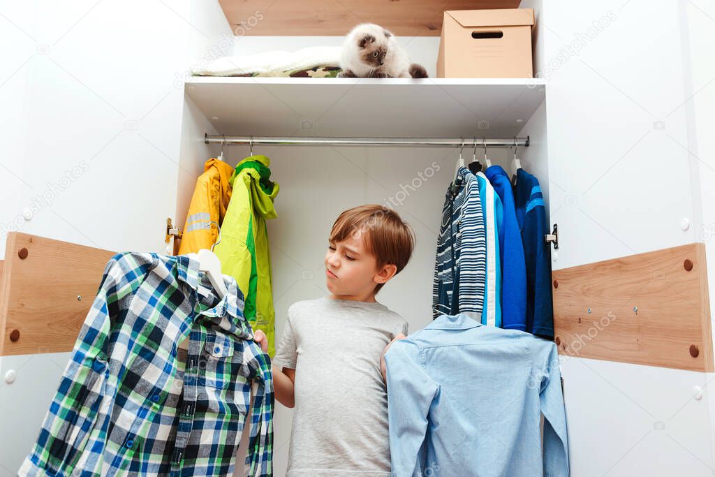 Young boy chooses clothes in the wardrobe closet at home. Cute boy taking school shirt to wear. Kid organizing clothes in wardrobe. Order in the closet. Wardrobe with child's clothing.