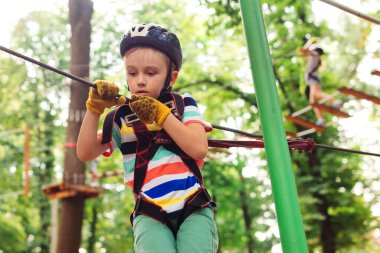 Macera parkında yüksek ip kursuna tırmanan sevimli çocuk. Kasklı çocuk, ekstrem spor. Çocuklar için yaz kampı. Çocuk ağaçların arasından kablo hattını geçiyor.