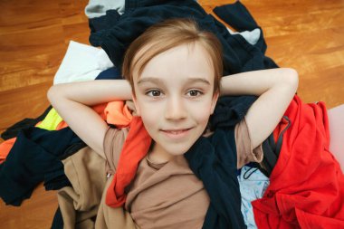 Dağınık kıyafetlerin arasında dağınık duran sevimli tembel çocuk portresi. Ev işleri ev işleri. Kirli çamaşırlarıyla yerde yatan dağınık çocuk..