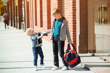 Okul konseptine geri dönelim. Sırt çantalı iki kardeş okula gidiyor. Ufaklık okula gitmek istemiyor. Arkadaşlar, çocukluk ve eğitim. İlkokul. Sokaklardaki moda çocukları..