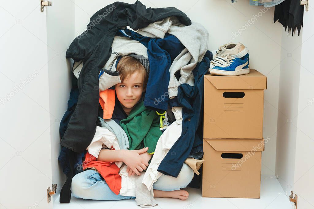 Kid hiding among messy clothes inside closet. Organization and storage of clothes at home. Mess in the wardrobe. Boy with messy colorful clothing at room. Untidy clutter clothing closet.