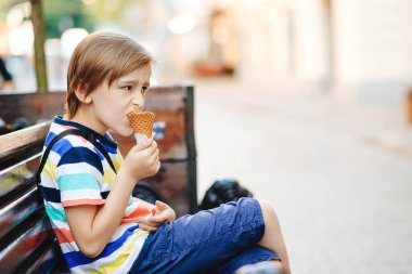Dondurma yiyen sevimli bir çocuk. Çocuk dışarıda bankta oturuyor. Hafta sonu yaz günü. Çocuk gevrek külahta lezzetli dondurmanın tadını çıkarıyor. Yaz tatili, yaşam tarzı ve moda konsepti.