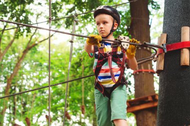 Macera parkında yüksek ip kursuna tırmanan sevimli çocuk. Kasklı çocuk, ekstrem spor. Çocuklar için yaz kampı. Çocuk ağaçların arasından kablo hattını geçiyor.