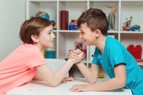 Cute boys competing in arm wrestling during the break time. Happy friends playing arm wrestle looking at each other. Cute brothers spending time together at home.