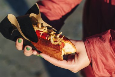 Taze sosisli sandviç ve ızgara sosis tutan bir kadın. Sandviç kağıdında servis edilir, yakın çekim. Sokak yemeği, fast food.