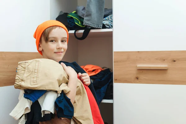 Boy standing near messy clothes on shelf. Preteen boy with dirty clothing in his room. Messy home kid's room. Untidy clutter clothing closet. Boy thinking what to do with messy.