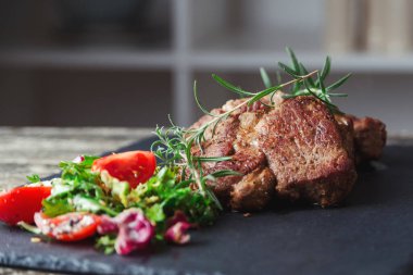 Siyah taş levhada ızgara biftek. Lezzetli ızgara biftek ve domatesli salata. Ana yemek sığır eti. Biftek ve salatalı akşam yemeği. Tahtada büyük para var..