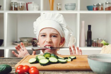 Ağzında bıçak olan kızgın bir aşçı. Çocuk profesyonel aşçı olmak istiyor. Çocuk şef salata için sebze doğruyor. Bıçaklı aşçı. Aşçı şapkası ve üniforması olan sevimli bir çocuk..