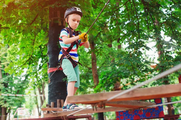 The boy climbs a rope park. Little cute boy in a climbing adventure park. Summer sunny day. Rope playground structure. Safe climbing extreme sport. Child with helmet insurance in a rope park.