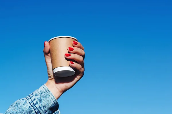 Woman hands holding a cup of coffee over blue sky background. Hipster girl holding paper cup with coffee take away. Paper coffee cup in woman hands with perfect manicure.