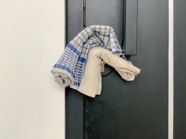 Siyah bir kapıya asılı ekose bir havlu ve bej bir havlu yakın çekim, kumaşların dokularını ve renklerini koyu arka planda sergiliyor..