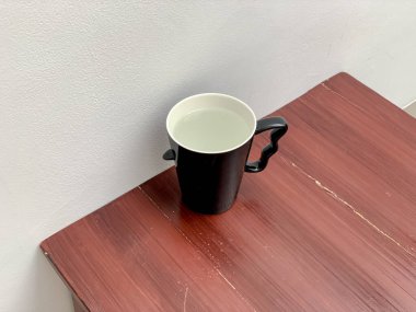 Basit bir siyah beyaz kupa, arka planında düz beyaz bir duvar olan kırmızımsı kahverengi bir tahta masanın üzerinde oturur. Görüntü, minimalist ayarı vurgulayarak, hafifçe yükseltilmiş bir açıdan alınır..