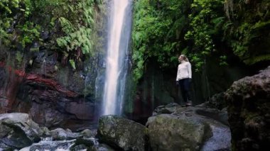 Kafkas yürüyüşçüsü, yüksek şelalenin altında duruyor, beyaz ceket, ıslak kayalarda sert botlar, derin düşüncelere dalma duruşu, sisli kanyon. Madeira, Portekiz