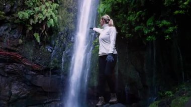 Kafkasyalı gezgin şelale sisine doğru uzanıyor, işaret ediyor, keşif jesti, ıslak bazalt adımları, tuhaf bir ifade. Madeira, Portekiz