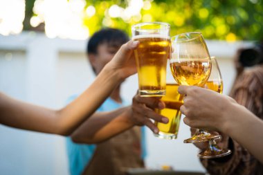 A group of multiracial friends are having a backyard dinner party, clinking beers or sharing drinks during happy hour. Young people are having fun together and clinking beers while standing in the garden.
