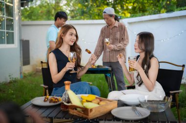 A group of multiracial friends are having a backyard dinner party, clinking beers or sharing drinks during happy hour. Young people are having fun together and clinking beers while standing in the garden.